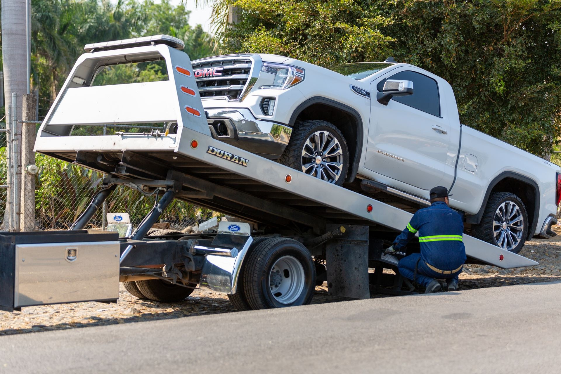 Grúa de plataforma con la rampa inclinada subiendo una camioneta blanca mientras un operario asegura el vehículo