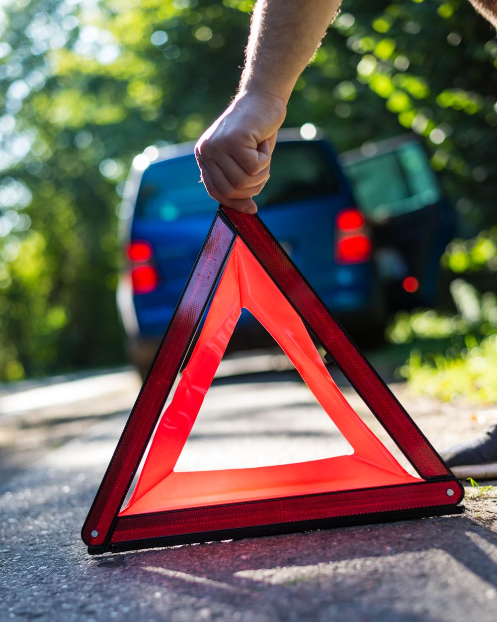 Mano colocando un triángulo de emergencia reflectante sobre el pavimento, con un auto detenido al fondo