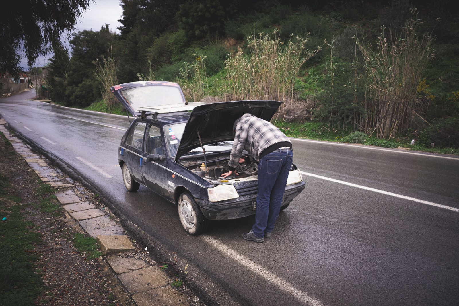 Conductor revisando el motor de su auto con el capó abierto, detenido en un camino mojado por la lluvia